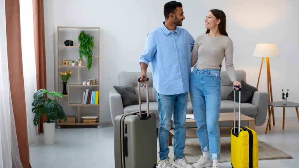 Casal abraçado e sorrindo em sala de estar com malas de viagem