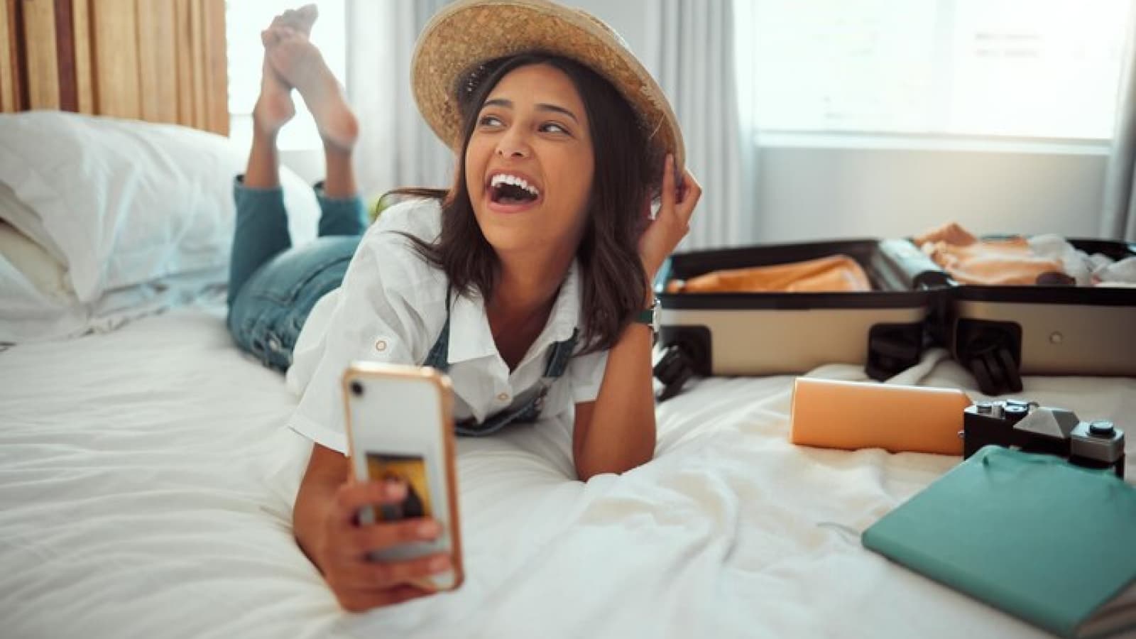 Mulher de chapéu deitada na cama com celular na mão e mala de viagem aberta